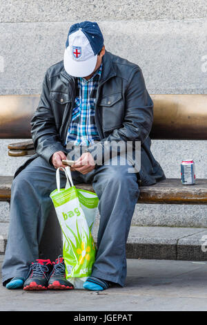 Un uomo di mezza età seduta su una panchina con le sue scarpe off, accanto ad una lattina di bevanda, guardando al suo telefono cellulare o telefono cellulare. Foto Stock