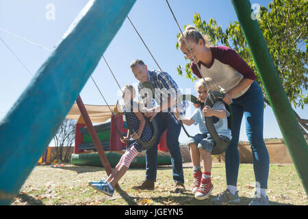 Genitori felici swinging bambini al parco giochi durante la giornata di sole Foto Stock