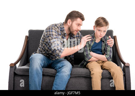 Padre abbracciando e parlando con sconvolto piccolo figlio seduto sul divano con bracci incrociati, problemi familiari concetto Foto Stock