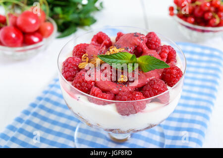 Dolce cremoso dessert con muesli, crema di formaggio, fresche Lamponi in una ciotola di vetro su un bianco sullo sfondo di legno. Close up Foto Stock