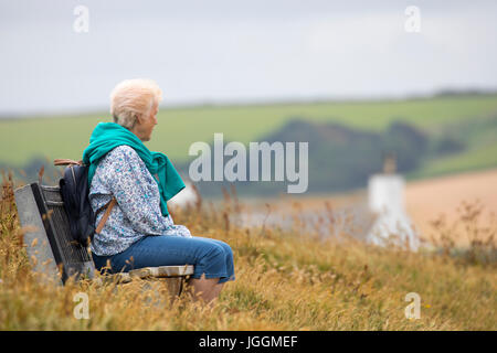 Un lone anziani di sesso femminile solo seduta su una panca in legno lungo il Cornish sentiero costiero tra Polzeath e Daymer Bay in Cornovaglia, Inghilterra cercando Foto Stock