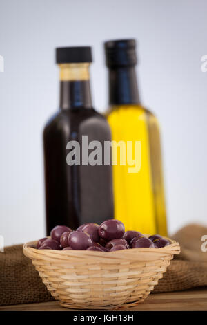 Close up di olive nel cesto di vimini con bottiglia di olio su tavola contro la parete Foto Stock