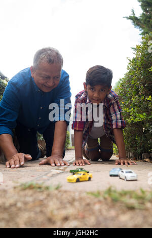 Ragazzo e nonno a giocare con modellini di auto in ginocchio sul marciapiede in cantiere contro sky Foto Stock