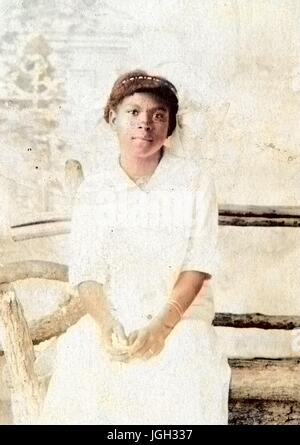 Tre quarti di ritratto di una giovane africana donna americana, seduto su una panca in legno, che indossa un abito bianco, indossando un anello di nozze, con un grave espressione facciale, 1920. Nota: l'immagine è stato colorizzato digitalmente usando un processo moderno. I colori possono non essere periodo-precisa. Foto Stock