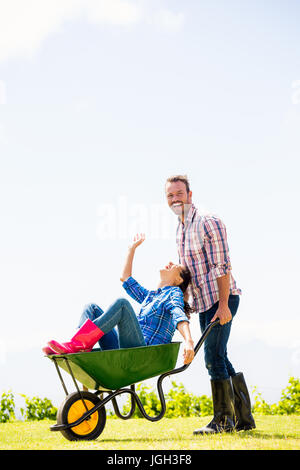 Felice l'uomo spinge donna seduta in carriola a prato Foto Stock