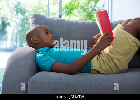 Vista laterale del ragazzo disteso durante la lettura del romanzo sul divano nel soggiorno di casa Foto Stock