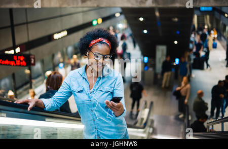 Elegante giovane donna africana ascolta musica sul suo cellulare durante la guida su una scala mobile in una trafficata Stazione della metropolitana Foto Stock