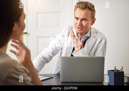 Giovani sorridente cosmetologo uomo seduto a un computer portatile in ufficio e comunicante con una donna. Foto Stock