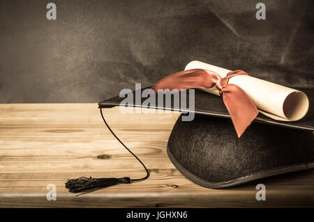 Un diploma scorrere legate con un nastro rosso, in appoggio su una graduazione mortarboard, sulla sommità di una scrivania in legno nella parte anteriore di una scuola di Blackboard. Creato in vinta Foto Stock