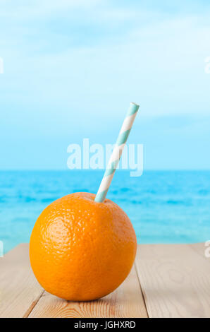 Una tutta arancione, perforata con una cannuccia a righe per rappresentare una fresca bevanda a base di frutta. Impostare su un planked tavolo in legno con soft focus il mare e il cielo dello sfondo. Foto Stock