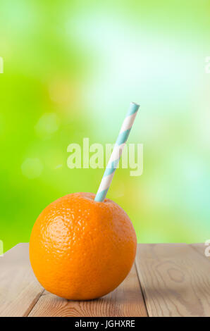 Tutta una frutta di arancia, perforata con una cannuccia a righe per rappresentare una fresca bevanda a base di frutta. Impostare su un planked tavolo in legno con soft focus giardino fogliame in Foto Stock