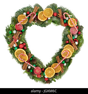 A forma di cuore ghirlanda di vischio con secchi frutta di arancia, mele, bastoncini di cannella, candy canes, ninnolo decorazioni, agrifoglio e inverno verde su bianco b Foto Stock