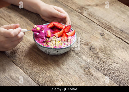 Mangiare sano colazione ciotola. Acai smoothie, granola, semi, fragole fresche in vaso di ceramica nella femmina (bambino) con le mani su sfondo di legno. Pulire Foto Stock