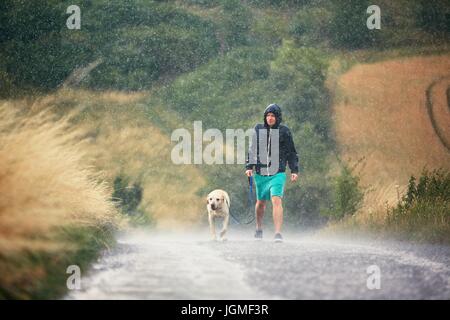 Giovane uomo che cammina con il suo cane (labrador retriever) in heavy rain per la strada rurale. Foto Stock