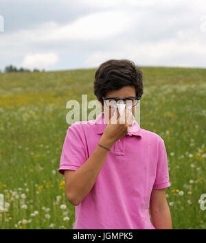 Allergia ragazzo con gli occhiali e t-shirt rosa soffia il naso con un fazzoletto bianco in primavera Foto Stock