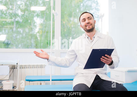 Allegro giovane medico tenendo un clipboard e gesticolando con la mano in ospedale Foto Stock