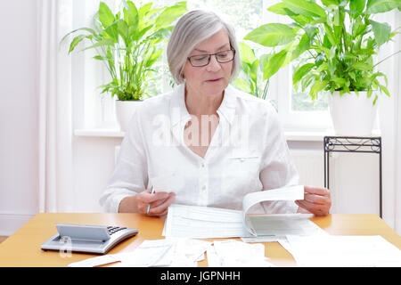 Senior donna tedesca a casa con una calcolatrice e lotti di fatture e ricevute, compilando i moduli stampati dell annuale dichiarazione fiscale Foto Stock