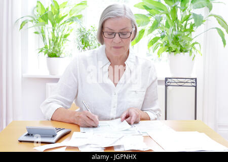 Senior donna tedesca a casa con una calcolatrice e lotti di fatture e ricevute, compilando i moduli stampati dell annuale dichiarazione fiscale Foto Stock