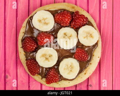 Pancake con cioccolato da spalmare con banana e lamponi contro un sfondo rosa Foto Stock