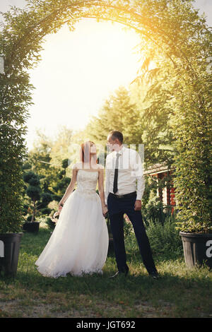 Bello lo sposo in camicia bianca e cravatta detiene la sposa la mano vicino a verde fiore archway con bel tramonto sullo sfondo Foto Stock