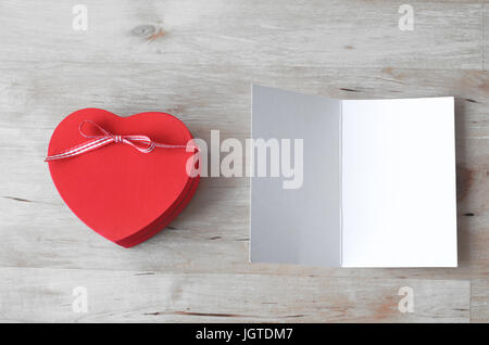 Tettuccio di colpo di un dipinto di rosso cuore di legno a forma di scatola regalo con nastro prua e una volta aperta la scheda Messaggi di saluto, lasciata vuota per un messaggio. Entrambi impostati su un paglierino Foto Stock