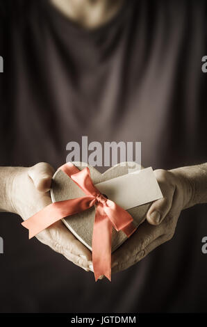 Maschio tenendo le mani su un cuore a forma di scatola regalo, legato ad un arco con nastro di raso. Un vuoto etichetta regalo facce anteriore per fornire spazio di copia per il messaggio. M Foto Stock