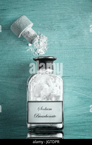 Un stile vintage bottiglia di bicarbonato di sodio (bicarbonato di sodio) sul lucido ciano riflettente o blu turchese superficie di legno. Cork aperto e polvere sparsi. Foto Stock