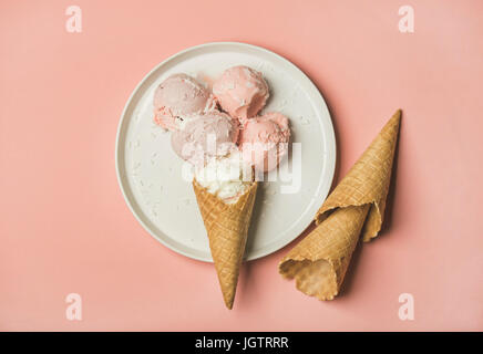 Flatlay di rosa pastello fragola e cocco gelato scoop e coni dolce sulla piastra bianca su rosa pastello, sfondo vista superiore Foto Stock