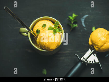 Estivo rinfrescante dessert. Mango sorbetto gelato scoop con menta fresca nella tazza verde nero su sfondo di legno, vista dall'alto. Pulire, mangiare sano, v Foto Stock