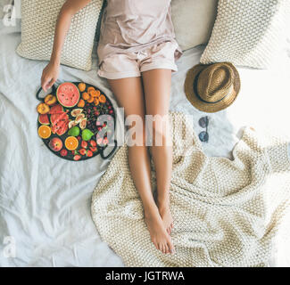 Estate sano materie vegan pulire mangiare la colazione a letto di concetto. Ragazza giovane indossando color pastello home vestiti prendendo frutto dal vassoio pieno di sé fresca Foto Stock