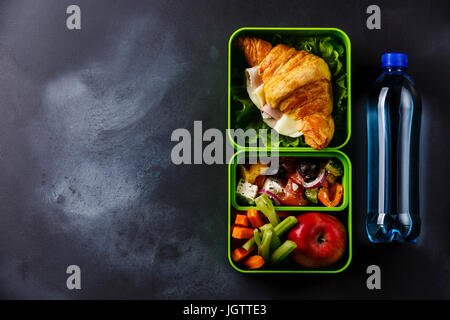 Estrarre il cibo scatola di pranzo con croissant sandwich, insalata greca e verdure con bottiglia di acqua sulla lavagna sfondo spazio di copia Foto Stock