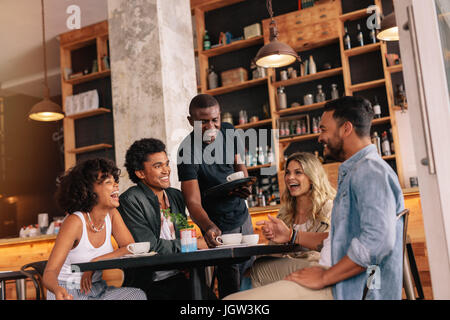 Gruppo di amici seduti attorno al tavolo e il cameriere che serve caffè presso il cafe. I giovani riuniti presso la caffetteria.