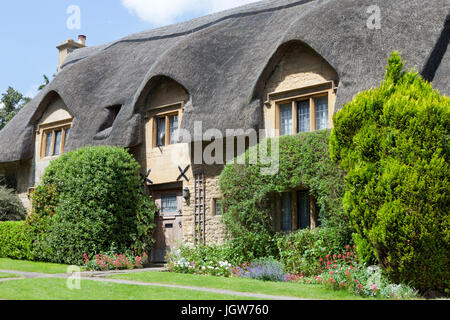 Tradizionale casa di pietra con il tetto di paglia e fiori, arbusti nel giardino anteriore, in un villaggio Inglese in Cotswolds, su una soleggiata giornata estiva . Foto Stock