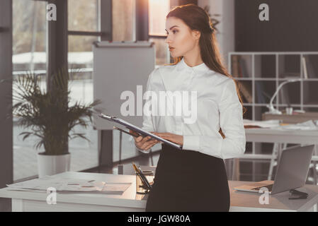 Attraente pensieroso imprenditrice holding negli appunti e in piedi in ufficio Foto Stock