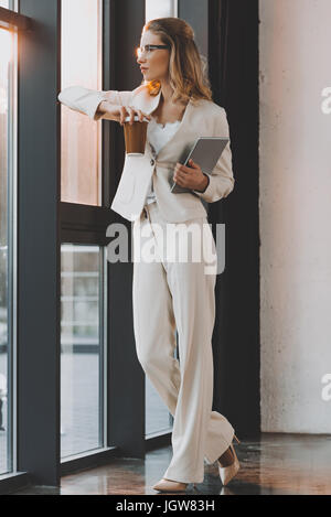 Attraente pensieroso imprenditrice in abito bianco con tavoletta digitale e caffè in piedi alla finestra in ufficio Foto Stock