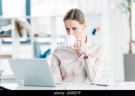 Giovane imprenditrice lavorando su laptop e di bere il caffè in ufficio Foto Stock