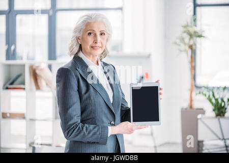 Sorridente imprenditrice senior in piedi in ufficio e azienda tavoletta digitale con schermo vuoto Foto Stock