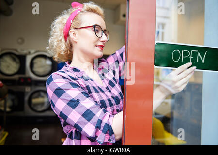 Femmina proprietario di piccola impresa mettendo a segno aperta sulla porta di una lavanderia a gettoni Foto Stock