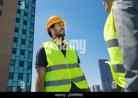 Due uomini che indossano copricapi rigidi e hi vis canottiere, avente la conversazione, all'aperto Foto Stock