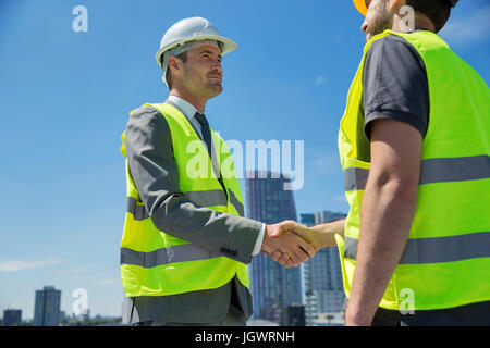 Due uomini che indossano copricapi rigidi e hi vis canottiere, stringono le mani, all'aperto Foto Stock