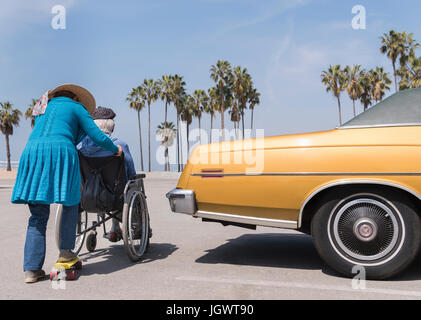 Senior donna spingendo il marito in carrozzella da vintage auto presso la spiaggia di Venice, California, Stati Uniti d'America Foto Stock