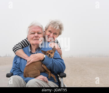 Ritratto di senior uomo in carrozzella con moglie e cane in spiaggia, Santa Monica, California, Stati Uniti d'America Foto Stock