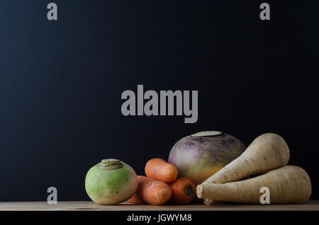 Ancora vita disposizione degli ortaggi a radice (rapa, carote, svedese e pastinaca) su un legno planked tabella con una lavagna in soft focus sfondo per Foto Stock