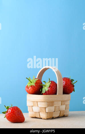 Rosso fresco Fragole, impilati per riempire un piccolo cesto tessuto su di una superficie di legno contro il fondo azzurro. Foto Stock