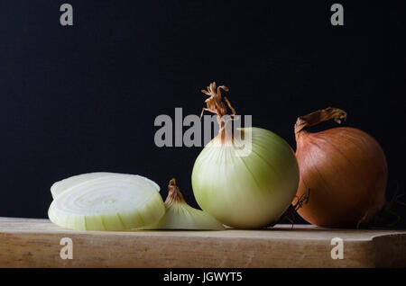 Ancora vita disposizione delle cipolle, interi, tagliati, pelati e non pelati su una cucina in legno tagliere contro uno sfondo nero. Foto Stock