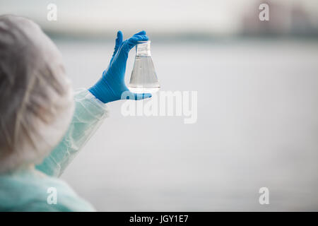 Foto di chimico dal retro ed in tuta protettiva con lampadina con acqua in mani sulla banca del fiume Foto Stock