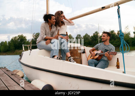 Tre amici rilassante ormeggiata sulla barca a vela, bere birra, l uomo a suonare la chitarra