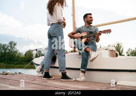Tre amici rilassante sul molo, seduti su una barca a vela, l'uomo a suonare la chitarra