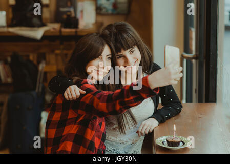 Due amiche, seduta in cafe, tenendo selfie, utilizza lo smartphone, tortina con candela sul contatore Foto Stock