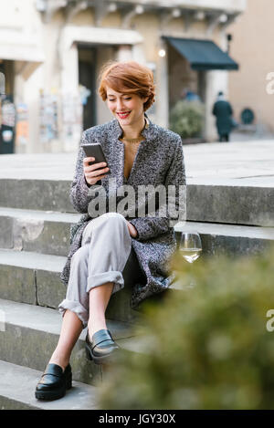 Giovane donna seduta sui gradini, utilizza lo smartphone, bicchiere di vino accanto a lei Foto Stock